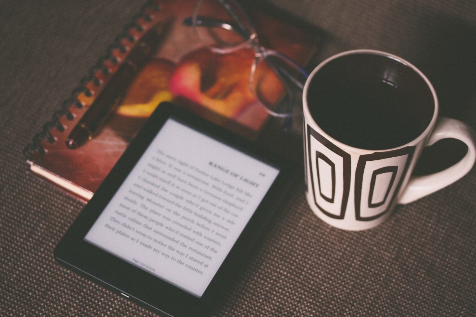 A kindle, notepad, and coffee mug on a brown table to show the main resources for upskilling and educating ourselves