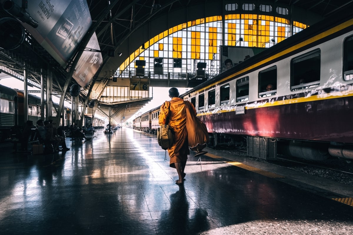 "a monk in yellow robe walks through Bangkok’s old train station to board his train"