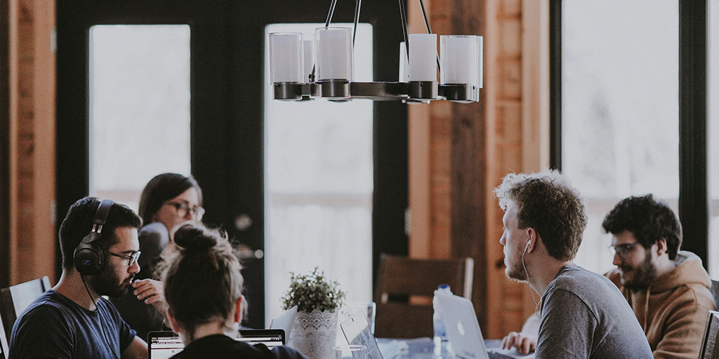 image of a group of people sitting on a rectangular table, some working with headphones on, some discussing