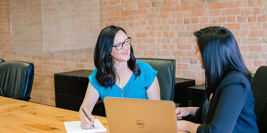 two women sitting on a desk talking with a laptop between them.